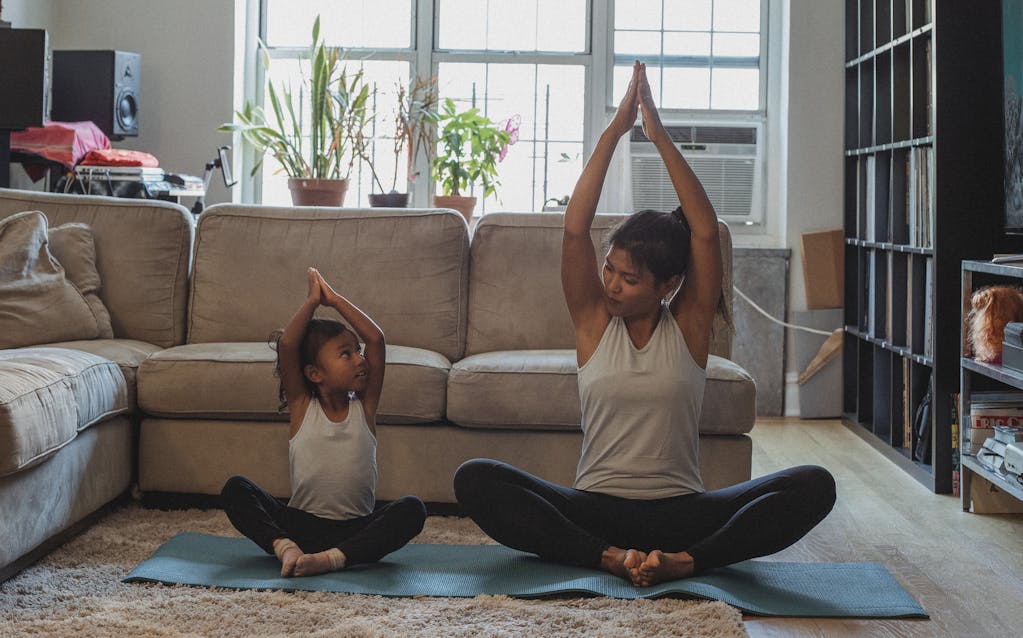 Full body content fit Asian woman and daughter in activewear sitting in Lotus Pose with arms raised and looking at each other while practicing yoga in cozy living room